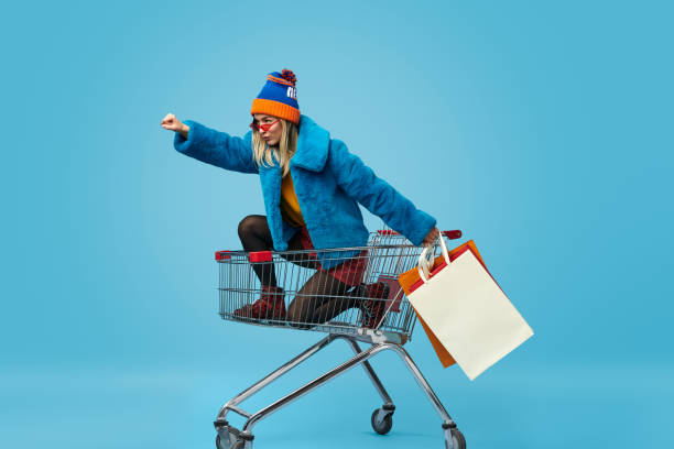 Side view of crazy trendy young female in bright clothes holding paper bags and riding cart in superhero pose against blue background